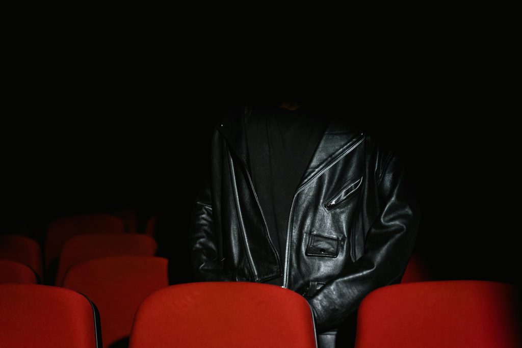 Individual wearing black leather jacket sits among red seats in a dimly lit cinema.